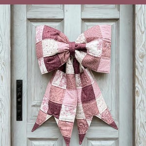 May include: A large, quilted bow in shades of pink and burgundy hangs on a white door. The bow is made of patchwork squares with floral and solid patterns. Text on the image reads "THE BIG GIANT QUILTED BOW" and "Designed to be reversible".