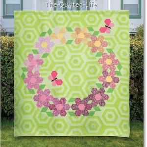 May include: A quilt with a lime green honeycomb pattern background, featuring a floral wreath design with pink, yellow, and orange flowers and two pink butterflies. The text "Flutterby Frolic" and "Krista Moser" are at the top. Sizes include Twin, Wall, and Table Runner.