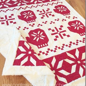 May include: A white and red quilted throw blanket with a snowflake and mitten pattern. The blanket is folded in half and has a white backing.