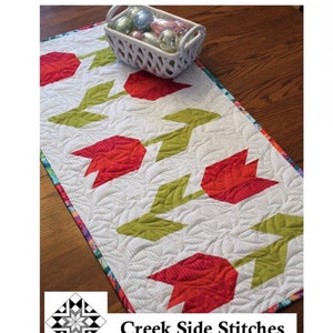 May include: A white table runner with a red and green tulip design. The runner is 16 inches by 37 inches. The text "Tulip Time" and "Creek Side Stitches #343" is printed on the image.