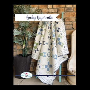 May include: A cream-colored quilt with a geometric pattern of blue and green shapes, including squares, triangles, and crosses. The quilt is draped over a light blue ladder. The text "Lucky Layercake" is displayed on a banner.
