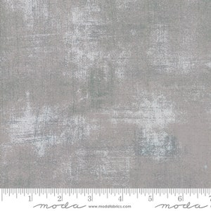 May include: A close-up of a gray fabric with a white and blue distressed pattern. The fabric has a subtle texture and a slightly faded look. The fabric is marked with a ruler that reads "moda" and "www.modafabrics.com".