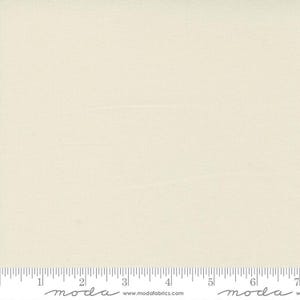 May include: Cream-colored fabric with a subtle texture, featuring a ruler along the bottom edge. The fabric appears to be a solid color, with a slight sheen. The word "moda" is printed on the ruler.