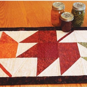 May include: A quilted table runner with a fall theme featuring three large maple leaves in shades of orange, red, and green. The leaves are made of fabric with a patchwork design. The runner has a black border.