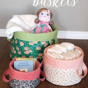 May include: Three fabric storage baskets with floral patterns. The largest basket is green with pink flowers and has a doll inside. The medium basket is pink with white flowers and is filled with white cloth. The smallest basket is gray with white flowers and has a book inside. The text "Olivia's baskets" is written above the baskets.