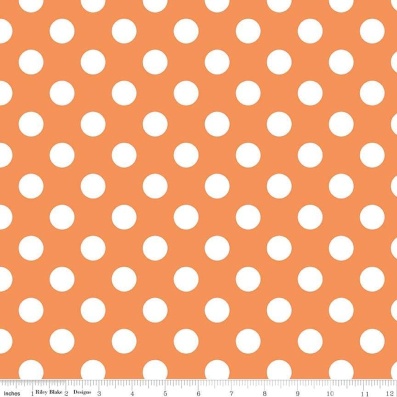 May include: Orange fabric featuring a repeating pattern of large white polka dots. A ruler with inch measurements is along the bottom edge. The text "Riley Blake Designs" is visible.