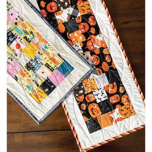 May include: A quilted table runner project with a Halloween theme. The runner features a patchwork design with orange pumpkins, black spiderwebs, and colorful squares. Finished size is 12 inches by 42 inches.