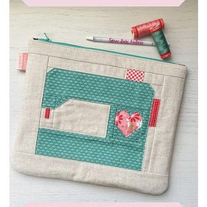 May include: A rectangular fabric pouch featuring a pieced design of a sewing machine in teal and beige with a pink heart accent. The pouch has a teal zipper, a small red and white tag, and is accompanied by a pen and two spools of thread.
