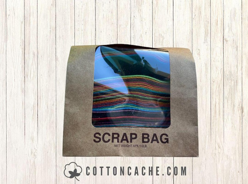 May include: A brown paper bag with a clear window displaying colorful fabric scraps. The bag is labeled "SCRAP BAG" with a net weight of approximately 1/2 lb. The website "cottoncache.com" is printed at the bottom.