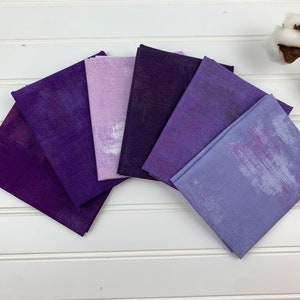 May include: A set of six purple and lavender fabric squares with a subtle brushed texture. The fabric squares are arranged in a gradient from dark purple to light lavender.