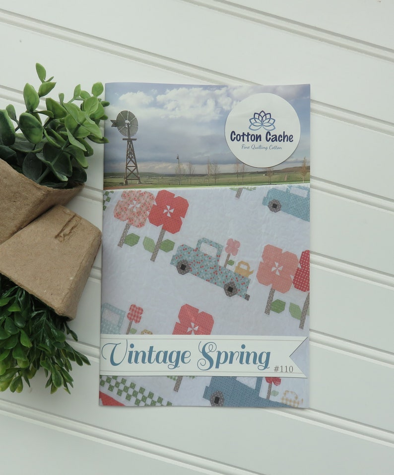 May include: A white fabric with a blue truck, pink flowers, and a windmill on a blue sky background. The fabric is labeled "Vintage Spring #110" and "Cotton Cache Fine Quilting Cotton".