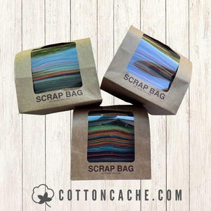 May include: Three brown paper bags labeled "SCRAP BAG" with a clear window showing colorful fabric scraps. The bags are arranged on a light wood-grain surface. The website address cottoncache.com is visible.