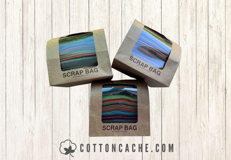 May include: Three brown paper bags labeled "SCRAP BAG" with a clear window showing colorful fabric scraps. The bags are arranged on a light wood-grain surface. The website address cottoncache.com is visible.