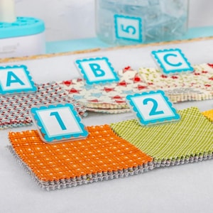 May include: A collection of colorful fabric squares with decorative edges, arranged in a row. Each square is paired with a small, square label featuring a number or letter (1, 2, A, B, C) in a blue scalloped frame. A jar with a label marked "5" is in the background.