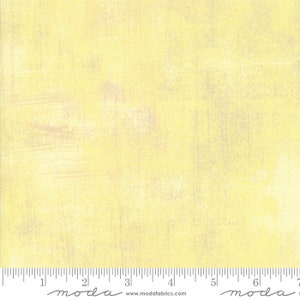 May include: A pale yellow fabric with a subtle, textured pattern. The fabric has a slightly distressed look, with hints of white and gray throughout. The fabric is marked with a ruler and the word "moda" in black.