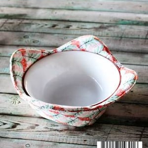 May include: A white bowl with a floral print fabric bowl cozy. The cozy has a red, green, and white floral pattern. The bowl and cozy are sitting on a wooden surface.