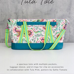 May include: A white tote bag with a floral print and teal accents. The bag has a luggage sleeve, d-rings, and multiple pockets. The bag is designed by Sallie Tomato in collaboration with Tula Pink.