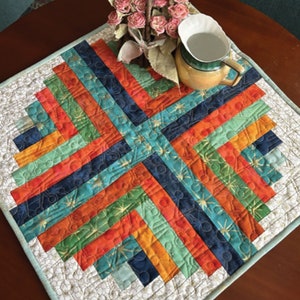 May include: A colorful quilted table runner with a geometric pattern in shades of orange, green, blue, and brown. The runner is quilted with a starburst design.