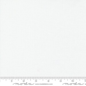 May include: White fabric with a subtle texture. The fabric is measured with a ruler that has the word "moda" printed on it.
