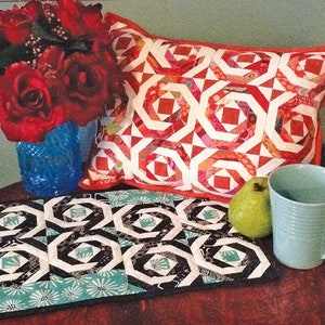 May include: A black and white geometric patterned table runner with a red and white geometric patterned pillow, a blue vase with red roses, a green pear, and a light blue mug.