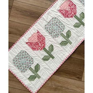 May include: A white table runner with a pink and green floral design. The runner is 16.5 inches by 42.5 inches and features a repeating pattern of stylized tulips. The text "Posy Patrol Table Runner" is at the top of the image. The text "by Chrissy Lux of Branch & Blume" is at the bottom of the image.