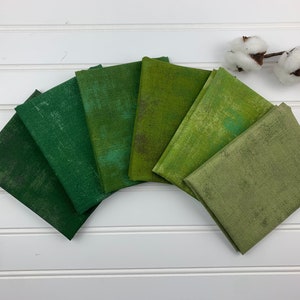 May include: Six fabric squares in various shades of green, arranged in a semi-circle on a white surface. The fabric has a subtle distressed texture.