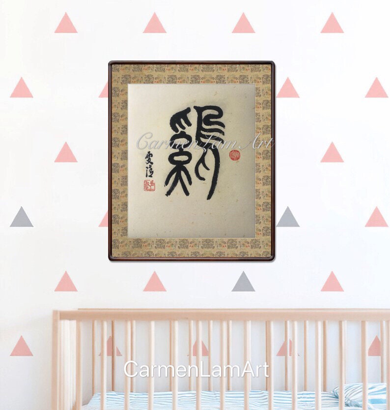 Puede incluir: Una obra de arte de caligraf&iacute;a enmarcada con tinta negra sobre un fondo beige. La obra presenta un solo car&aacute;cter chino en estilo tradicional. El marco es de madera marr&oacute;n con un borde dorado. La obra est&aacute; colgada en una pared blanca con pegatinas triangulares rosas y grises.
