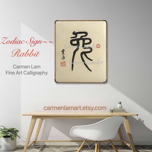 Könnte beinhalten: Ein Schwarzweiß-Kalligraphie-Gemälde des chinesischen Zeichens für "Kaninchen" auf beigefarbenem Hintergrund. Das Gemälde ist in einem Holzrahmen gerahmt. Der Name des Künstlers, "Carmen Lam Art", ist in roter Tinte in der unteren rechten Ecke des Gemäldes geschrieben.
