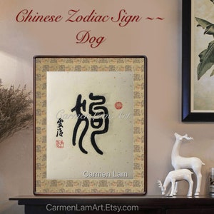 Puede incluir: Una obra de arte de caligraf&iacute;a enmarcada que presenta el car&aacute;cter chino para "perro" en tinta negra sobre un fondo beige. La obra de arte se titula "Signo del zod&iacute;aco chino - Perro" y est&aacute; firmada por "Carmen Lam".