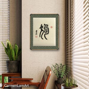 Puede incluir: Una obra de arte de caligraf&iacute;a enmarcada que presenta el car&aacute;cter chino "龍" (long) que significa drag&oacute;n. La obra de arte est&aacute; sobre un fondo beige con un marco verde y un sello rojo.