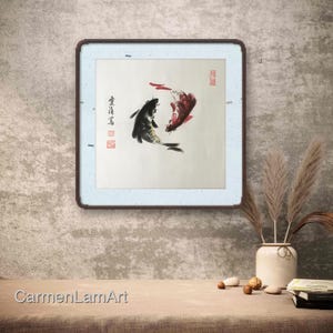 May include: A framed artwork featuring two koi fish in a traditional Asian ink painting style. One fish is black, the other red, swimming in a circular pattern. The artwork is in a square frame with a light blue border and a dark brown frame.