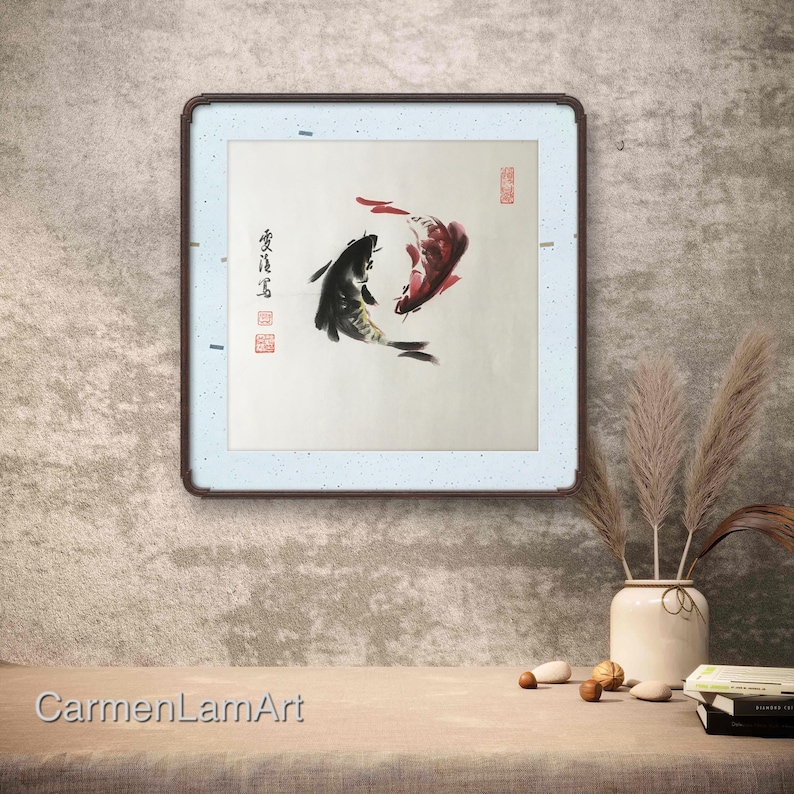 May include: A framed artwork featuring two koi fish in a traditional Asian ink painting style. One fish is black, the other red, swimming in a circular pattern. The artwork is in a square frame with a light blue border and a dark brown frame.