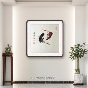 May include: A framed artwork featuring a traditional Asian ink painting of two koi fish, one black and one red, swimming in opposite directions. The artwork is set against a white background and framed in a dark wood frame. The artist's name is at the bottom.