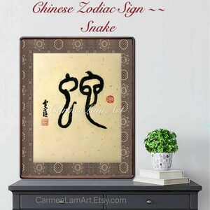 Pode incluir: Obra de arte emoldurada com um símbolo de caligrafia chinesa preta representando o signo do zodíaco da Serpente. A obra de arte tem um fundo bege e uma borda marrom estampada. O texto "Chinese Zodiac Sign ~ Snake" está no topo.