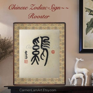 Puede incluir: Una pintura china de caligrafía enmarcada que representa el signo del zodíaco chino del Gallo. La pintura está sobre un fondo crema con tinta negra. Los caracteres chinos para Gallo están escritos en caligrafía china tradicional. La pintura está enmarcada en un marco de madera oscura con un borde dorado.