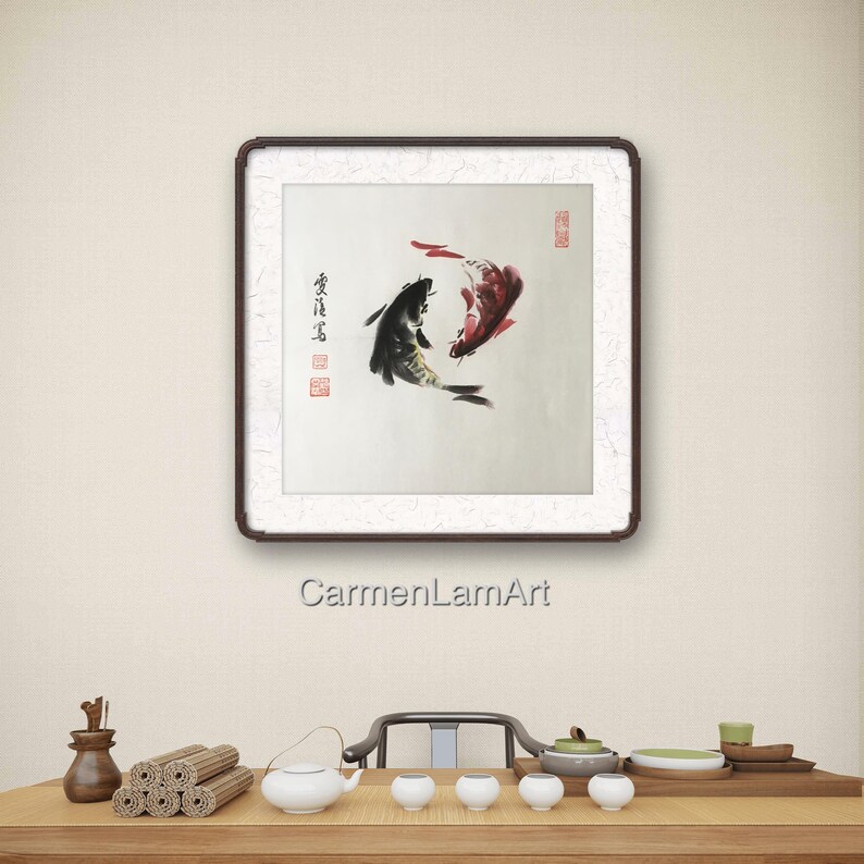 May include: Square framed artwork featuring two koi fish in a traditional Asian ink painting style. One fish is black, the other red, set against a white background. The frame is dark brown. The artwork is displayed above a wooden table.