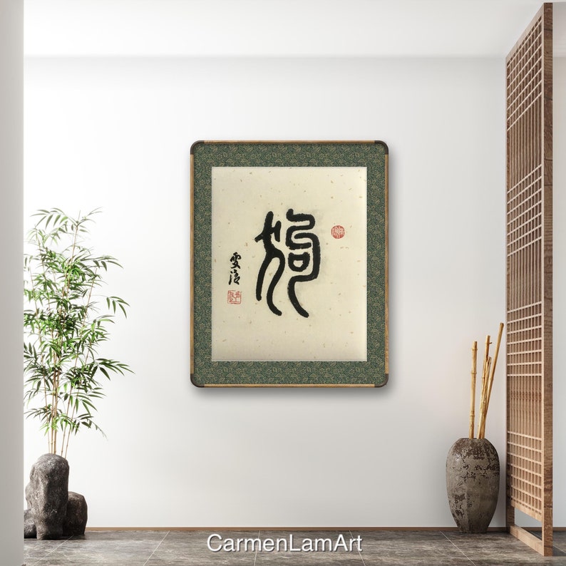 Puede incluir: Una obra de arte de caligraf&iacute;a enmarcada que presenta el car&aacute;cter chino "龍" (long) en tinta negra sobre un fondo beige. El car&aacute;cter est&aacute; rodeado por un borde verde y un sello rojo. La obra se exhibe en una pared blanca en un entorno interior moderno.