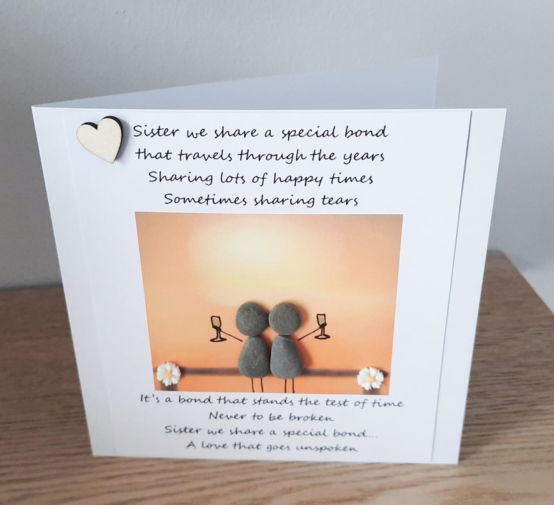 May include: A card with a painted image of two figures made of stones holding glasses of champagne. The card has a poem about sisterhood that reads "Sister we share a special bond that travels through the years. Sharing lots of happy times. Sometimes sharing tears. It's a bond that stands the test of time. Never to be broken. Sister we share a special bond... A love that goes unspoken."