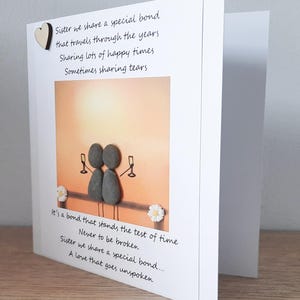 May include: A card featuring a pebble art illustration of two figures raising a toast with glasses of champagne. The card includes a poem about sisterhood that reads "Sister we share a special bond that travels through the years Sharing lots of happy times Sometimes sharing tears It's a bond that stands the test of time Never to be broken Sister we share a special bond... A love that goes unspoken"