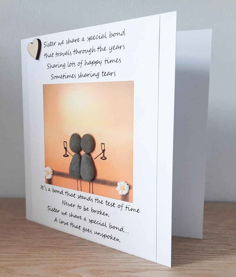 May include: A card featuring a pebble art illustration of two figures raising a toast with glasses of champagne. The card includes a poem about sisterhood that reads "Sister we share a special bond that travels through the years Sharing lots of happy times Sometimes sharing tears It's a bond that stands the test of time Never to be broken Sister we share a special bond... A love that goes unspoken"