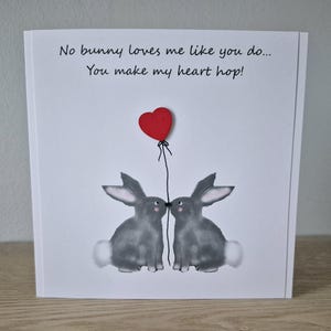 Może przedstawiać: Biała kartka z czerwonym balonem w kształcie serca i dwoma szarymi kr&oacute;likami, kt&oacute;re się całują. Na kartce napisano: "No bunny loves me like you do... You make my heart hop!"
