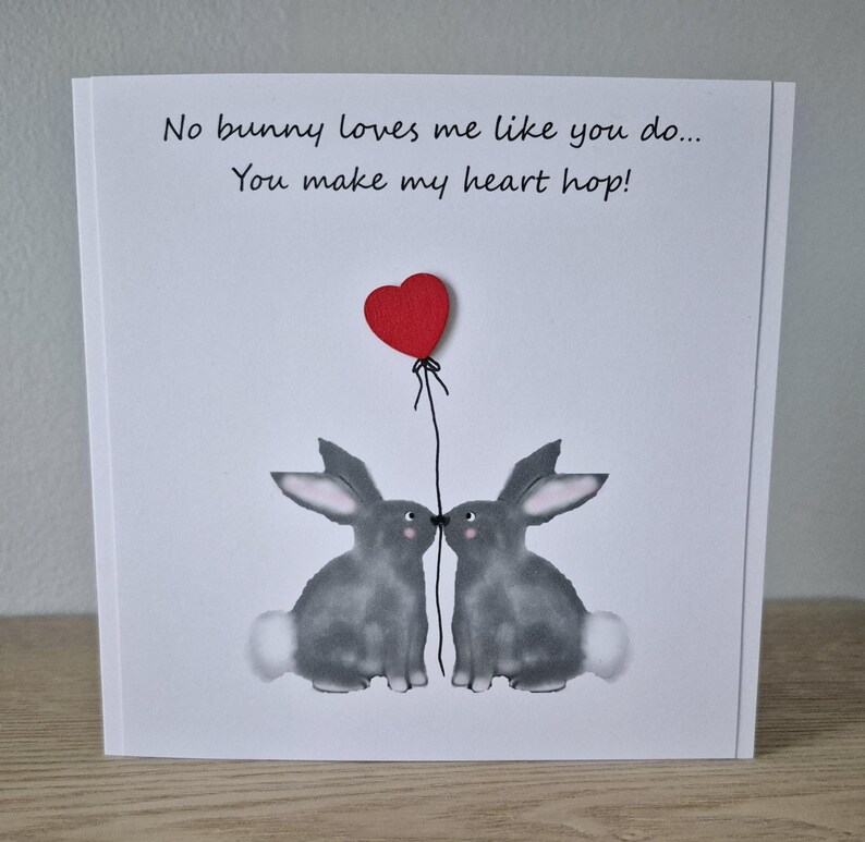 Może przedstawiać: Biała kartka z czerwonym balonem w kształcie serca i dwoma szarymi kr&oacute;likami, kt&oacute;re się całują. Na kartce napisano: "No bunny loves me like you do... You make my heart hop!"