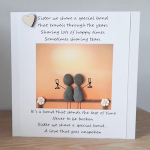 May include: A card featuring a hand-painted pebble art design of two figures holding glasses of champagne. The card has a message about sisterhood and a special bond that travels through the years. The text reads: "Sister we share a special bond that travels through the years Sharing lots of happy times Sometimes sharing tears. It's a bond that stands the test of time Never to be broken Sister we share a special bond... A love that goes unspoken"