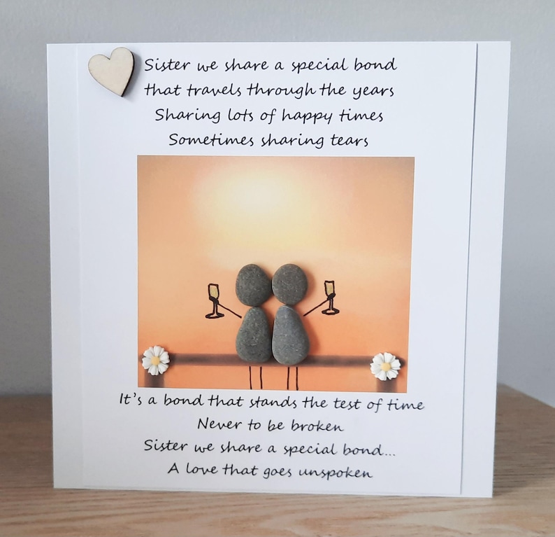 May include: A card featuring a hand-painted pebble art design of two figures holding glasses of champagne. The card has a message about sisterhood and a special bond that travels through the years. The text reads: "Sister we share a special bond that travels through the years Sharing lots of happy times Sometimes sharing tears. It's a bond that stands the test of time Never to be broken Sister we share a special bond... A love that goes unspoken"