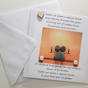 May include: A card with a handwritten message about sisterhood. The card features a sunset background with two figures made of stones holding glasses of wine. The text reads "Sister we share a special bond that travels through the years Sharing lots of happy times Sometimes sharing tears. It's a bond that stands the test of time Never to be broken Sister we share a special bond..... A love that goes unspoken"