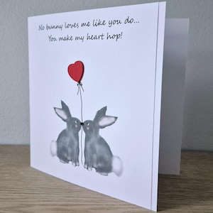 Może przedstawiać: Kartka z życzeniami z dwoma szarymi kr&oacute;likami całującymi się pod czerwonym balonem w kształcie serca. Na kartce napisano "No bunny loves me like you do... You make my heart hop!"