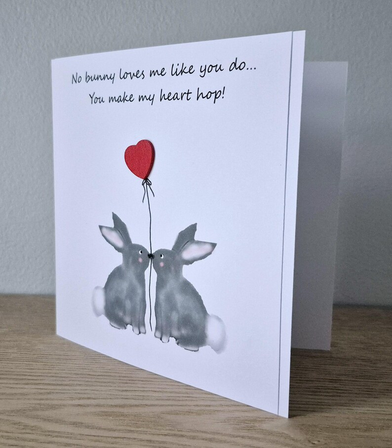 Może przedstawiać: Kartka z życzeniami z dwoma szarymi kr&oacute;likami całującymi się pod czerwonym balonem w kształcie serca. Na kartce napisano "No bunny loves me like you do... You make my heart hop!"