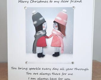 May include: A Christmas card with a winter scene featuring two figures holding hands. The figures wear hats, scarves, and coats in shades of pink, red, and gray. Text reads "Merry Christmas to my dear friend" and a message of friendship.