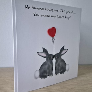 Może przedstawiać: Biała kartka z ręcznie napisanym czarnym tekstem: "No bunny loves me like you do... You make my heart hop!" Na kartce narysowane są dwa szare kr&oacute;liki, kt&oacute;re patrzą na siebie. Nad kr&oacute;likami przymocowane jest czerwone serce do sznurka.