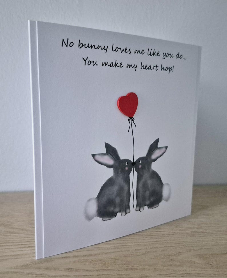 Może przedstawiać: Biała kartka z ręcznie napisanym czarnym tekstem: "No bunny loves me like you do... You make my heart hop!" Na kartce narysowane są dwa szare kr&oacute;liki, kt&oacute;re patrzą na siebie. Nad kr&oacute;likami przymocowane jest czerwone serce do sznurka.